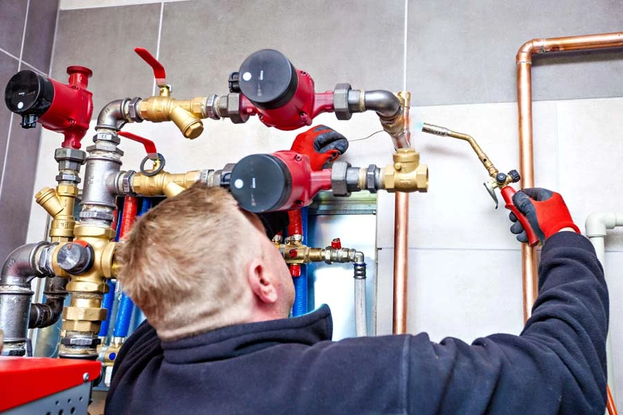 Plombier professionnel travaillant sur une installation de chauffage en intérieur, manipulant des outils sur un système complexe de tuyauterie cuivre et raccords