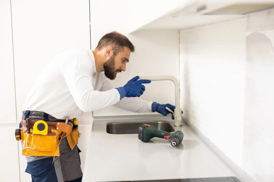 Un plombier professionnel en tenue blanche et gants de protection bleus répare un robinet de cuisine dans un intérieur moderne